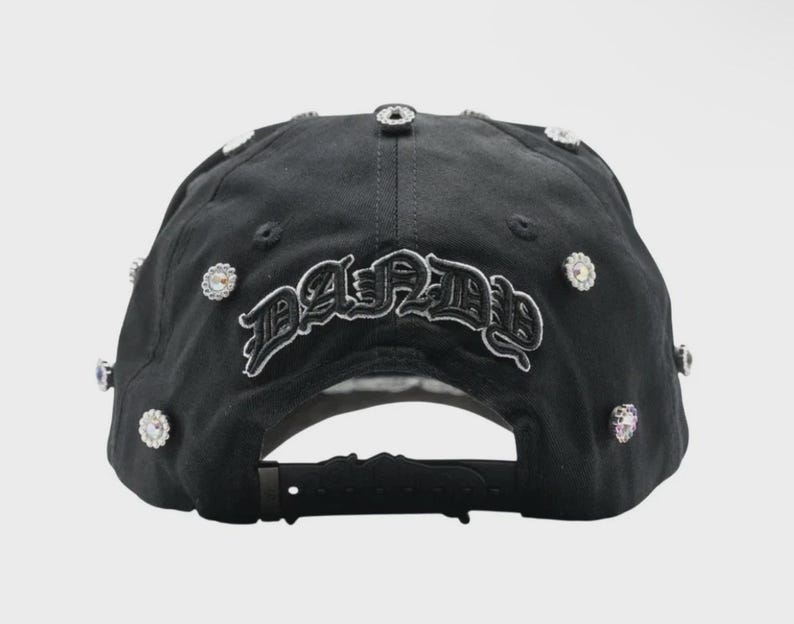 May include: Black baseball cap with the word "PARIS" embroidered in a gothic font. The cap is adorned with multiple clear, sparkling rhinestones. The adjustable strap is visible at the back.