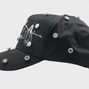 May include: Black baseball cap adorned with sparkling, jewel-like embellishments. The cap features white text and a curved brim. The phrase "it's me" is printed on the brim. A stylish accessory.