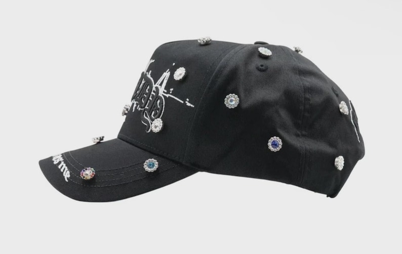 May include: Black baseball cap adorned with sparkling, jewel-like embellishments. The cap features white text and a curved brim. The phrase "it's me" is printed on the brim. A stylish accessory.