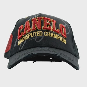 May include: Black baseball cap with the word "CANELO" in red and gold embroidery. The text "UNDISPUTED CHAMPION" is embroidered in gold. The cap has a distressed look and a red patch on the side.