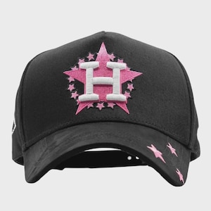 May include: Black baseball cap with a pink and white embroidered star design featuring the letter "H". Pink star accents are on the brim. A stylish accessory for sports fans.