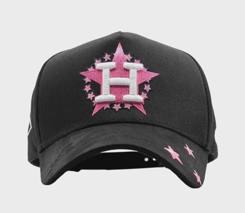 May include: Black baseball cap with a pink and white embroidered star design featuring the letter "H". Pink star accents are on the brim. A stylish accessory for sports fans.