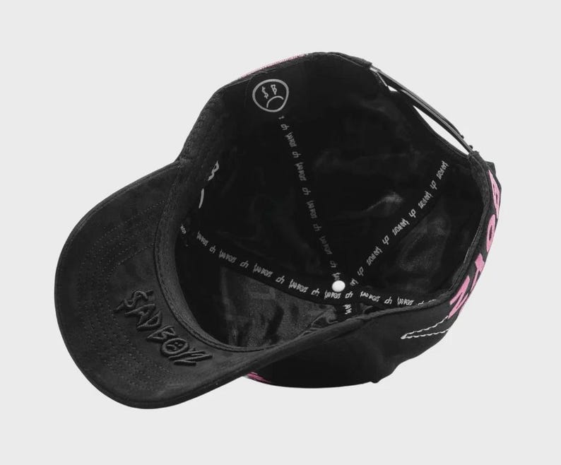 May include: Black baseball cap with a curved brim and adjustable strap. The cap features the word "SADBOY" embroidered on the front and pink text on the side. The interior has a white logo and text.