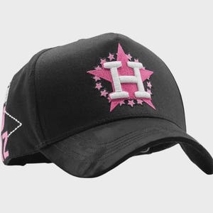 May include: Black baseball cap with a pink star and the letter "H" embroidered on the front. The star is surrounded by smaller pink stars. The cap has a curved brim and a structured crown. The side of the cap has a white and pink design.