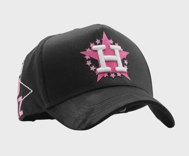 May include: Black baseball cap with a pink star and the letter "H" embroidered on the front. The star is surrounded by smaller pink stars. The cap has a curved brim and a structured crown. The side of the cap has a white and pink design.