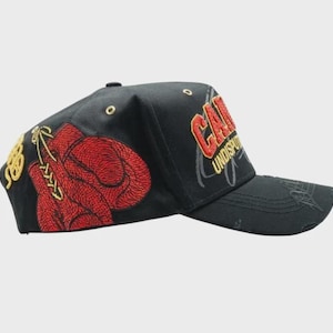 May include: Black baseball cap with embroidered red boxing gloves and gold accents. The cap features the word "CAN" in red and gold embroidery, with "UNDISPUTED" below. A stylish accessory for sports enthusiasts.
