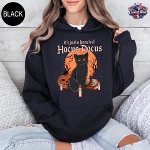 May include: A black hooded sweatshirt featuring a graphic print of a black cat sitting in front of an orange moon, a pumpkin, and a stack of books. The text reads, "it's just a bunch of Hocus Pocus." The design includes lit candles and silhouettes of bare trees.