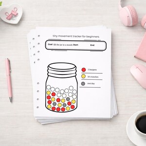 May include: A stack of white paper sheets with a fitness tracker design. The tracker is a jar graphic with red, yellow, and gray circles, and the text "tiny movement tracker for beginners". A pink pen, headphones, and a coffee cup are also visible.