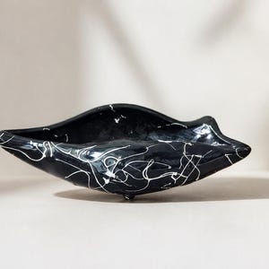 Peut inclure: Un bol en céramique noir de forme asymétrique. Le bol est orné de gribouillis blancs abstraits. Il repose sur trois petits pieds et convient à un usage décoratif.