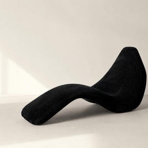 May include: A modern, black chaise lounge with a curved, flowing design. The chair is made of a soft, textured material and is set against a neutral background. The design is minimalist and elegant, suitable for a contemporary living space.