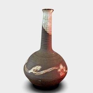 Peut inclure: Un vase en céramique fait à la main, avec un corps arrondi et un long col fin. Le vase est principalement brun foncé avec une surface texturée et présente un motif floral en crème et rouge. Le col est clair avec un bord marron.