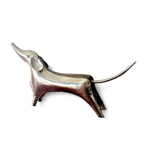 May include: A silver-colored metal sculpture of a dachshund dog in a stylized pose. The dog is depicted with an elongated body, short legs, and a long tail. The sculpture has a smooth, reflective surface and is set against a white background.