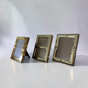 Peut inclure: Trois cadres photo en métal doré poli disposés en rangée. Le premier est un rectangle vertical, le deuxième est un cadre vertical à deux ouvertures superposées, et le troisième est un cadre carré avec des détails décoratifs aux coins.