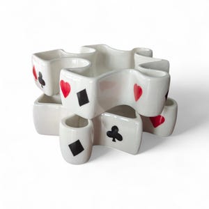 May include: White ceramic playing card-themed container with multiple compartments. Each compartment features a different playing card suit in red and black. The container has a unique, interlocking design.