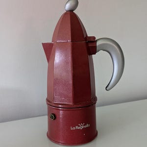 May include: A maroon moka pot coffee maker with a metallic finish, featuring a silver-coloured handle and a matching silver knob on the lid. The base of the coffee maker displays the white text La Reginella with a small crown icon above the name.