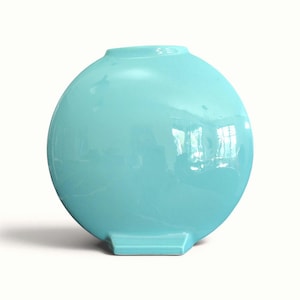 May include: A circular, turquoise ceramic vase with a smooth, glossy finish. The vase features a flat, rectangular base that supports the rounded body, which has a small, narrow opening at the top centre for holding items or floral arrangements.