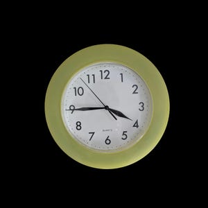 May include: A round wall clock with a pale green frame and a white face. The clock has black hands and black numerals. The word "QUARTZ" is printed at the bottom of the clock face.