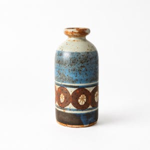 Peut inclure: Un vase en céramique avec un corps cylindrique et un col étroit. Le vase présente des bandes de couleurs, dont le bleu, le marron et le blanc. Le motif comprend des cercles bruns répétés avec un motif en forme d'étoile au centre. Le vase a une texture tachetée.