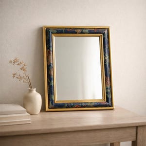 Peut inclure: Un miroir rectangulaire avec un cadre doré et une bordure à motifs floraux dans les tons de bleu, rouge et vert. Le miroir est posé contre un mur blanc sur une table en bois. Un petit vase blanc avec des fleurs séchées est à proximité.