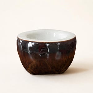 Peut inclure: Un petit bol en céramique fait à la main, avec un extérieur brun foncé et un intérieur blanc. Le bol a une forme arrondie avec un bord légèrement évasé. La finition brillante reflète la lumière, soulignant les variations de couleur riches.