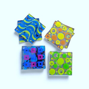 Peut inclure: Quatre dessous de verre carrés en verre avec des motifs géométriques colorés. L'un présente des lignes ondulées bleues et jaunes, un autre des cercles orange et jaunes, un troisième des carrés bleus et des rectangles roses, et le dernier des cercles verts.