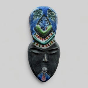 Peut inclure: Un masque allongé artisanal avec un visage noir et une coiffe bleue décorée. La coiffe présente des accents verts, rouges et blancs. Le masque a les yeux fermés et un design détaillé, probablement pour la décoration murale ou l'exposition culturelle.