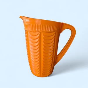 May include: An orange plastic pitcher with a handle and a spout. The pitcher has a textured design of vertical lines and curved shapes. The pitcher is empty and sits against a light blue background.