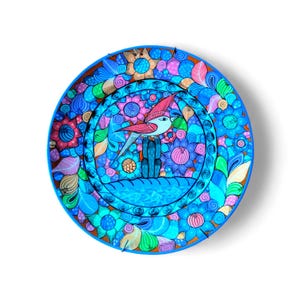 Peut inclure: Une assiette décorative ronde peinte à la main avec un motif vibrant d'oiseaux et de fleurs. L'assiette présente un bord bleu et une image centrale d'un oiseau perché sur un cactus, entouré de fleurs et de feuilles colorées. La palette de couleurs comprend des nuances de bleu, de rose et de vert.