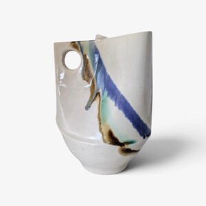 May include: A ceramic pitcher with an off-white glazed finish featuring a prominent side handle with a circular cutout. A diagonal streak of blue, green, and brown glaze runs across the body, creating a distinct contrast against the white surface.