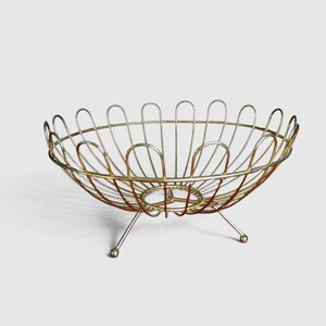 Peut inclure: Une corbeille à fruits en fil métallique doré présentant un design ajouré large avec une série de boucles arrondies en forme de pétales formant le bord. La corbeille est soutenue par trois pieds métalliques fins et inclinés, terminés par une petite boule.