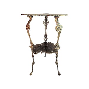 May include: A three-legged metal plant stand featuring a dark bronze finish with gold-toned highlights. The stand has an ornate, openwork design with curved legs and a triangular lower shelf, supporting a flat, decorative top surface.