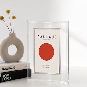 Peut inclure: Cadre photo en acrylique transparent avec bordure argentée, affichant une affiche de l'exposition Bauhaus avec un cercle rouge. Le cadre est incurvé en haut et en bas. Un petit vase avec des fleurs jaunes et des livres en arrière-plan.