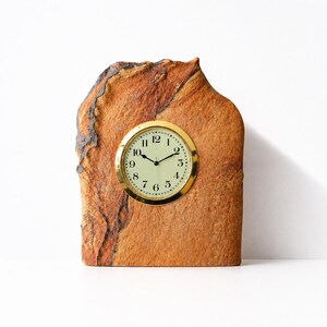May include: A decorative desk clock with a round gold-tone clock face set into a natural stone base. The clock face has black numerals and hands on a cream-colored background. The stone is a mix of brown and tan.