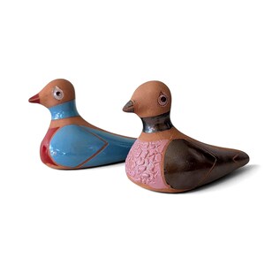 Peut inclure: Deux figurines d'oiseaux en céramique. L'oiseau de gauche a un corps bleu clair avec des accents rouges et une tête beige. L'oiseau de droite a une poitrine rose texturée, une aile brun foncé et une tête beige. Ils ont de petits yeux ronds.