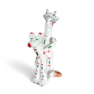 Peut inclure: Une sculpture en céramique blanche représentant deux personnages enlacés. Les personnages sont décorés de tirets rouges et verts. L'un des personnages a un couvre-chef en forme de couronne et l'autre une tête de chien. La base est de couleur orange clair.