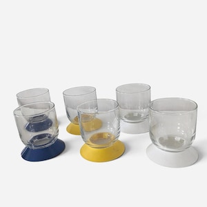 Può includere: Un set di sei bicchieri in vetro trasparente, ciascuno dotato di un'ampia base circolare di colore solido. La collezione comprende due bicchieri con base blu navy, due con base gialla e due con base bianca, disposti su una superficie piana.