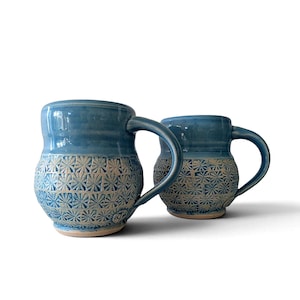 Peut inclure: Deux tasses en céramique avec un vernis bleu brillant sur la partie supérieure et une base texturée de couleur claire ornée d'un motif répétitif en forme d'étoile ou de fleur. Chaque tasse possède une anse courbée et un corps arrondi.
