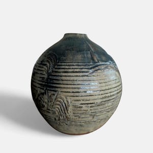 May include: A handmade ceramic vase with a rounded form and a narrow neck. The vase has horizontal, textured lines in a light beige colour, contrasted by dark grey accents. The design includes a textured, organic pattern on the side.