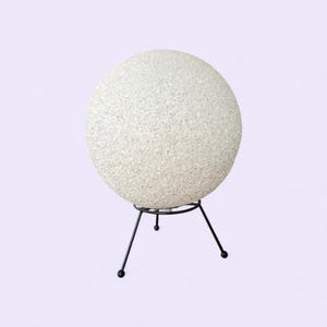 May include: A white, textured globe lamp with a black metal tripod base. The globe has a speckled appearance, and the lamp is set against a light purple background. The lamp is a decorative lighting fixture.