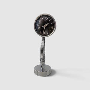 May include: A silver-toned desk clock featuring a circular face with a dark, reflective dial, white numerals at the 3, 6, 9, and 12 positions, and thin silver hands. The clock is supported by a flexible metal coil stem atop a circular base.