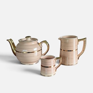 Peut inclure: Un service à thé de trois pièces comprenant une théière, un pot à lait et un pichet, tous en céramique rose pâle avec des rayures horizontales dorées et des anses et becs assortis, offrant une apparence coordonnée et raffinée.
