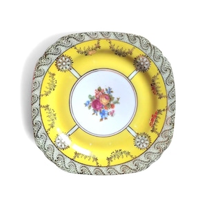 Peut inclure: Une assiette carrée en céramique avec une bordure jaune vif ornée de motifs en filigrane doré et quatre médaillons floraux circulaires. Le centre est blanc avec un bouquet coloré de roses et de petites fleurs bleues, bordé d'un contour festonné.