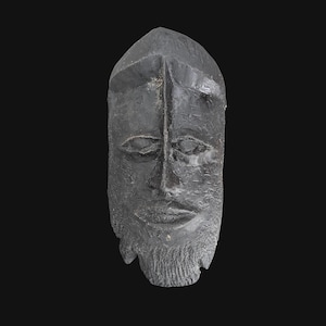 May include: A dark gray, carved wooden mask with a textured surface. The mask features a stylized face with closed eyes, a prominent nose, and a beard. The mask has a pointed top and is set against a black background.