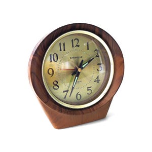 May include: A Caravelle table clock with a circular gold-toned face, black Arabic numerals, and a red second hand. The clock is encased in a smooth, dark brown wood frame with a distinct, flared base design and a clear protective glass cover.