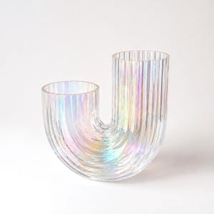 Peut inclure: Un vase en verre transparent et irisé avec un design en forme de U unique. Le vase présente des détails verticaux côtelés et un éclat arc-en-ciel, reflétant la lumière. Il possède deux ouvertures. Un objet de décoration intérieure.
