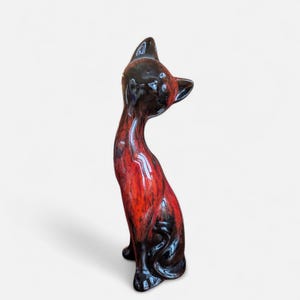 Peut inclure: Figurine de chat en céramique, en position assise, avec une finition brillante. Le corps du chat est rouge vif, passant au noir à la base et à la tête. Le chat regarde vers le haut, mettant en valeur sa forme élégante et allongée.