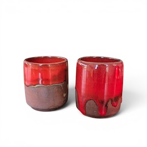 Peut inclure: Deux tasses en céramique faites à la main. Les tasses sont cylindriques avec un glaçage bicolore. La moitié supérieure est d'un rouge vif brillant, et la moitié inférieure est d'un brun terreux mat. L'émail rouge coule sur les côtés d'une tasse.