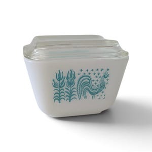 Peut inclure: Un plat de conservation en verre opalin blanc avec un couvercle en verre transparent. L'extérieur présente un motif graphique bleu sarcelle représentant deux épis de maïs et un coq entourés de petites formes en étoile. Forme rectangulaire.