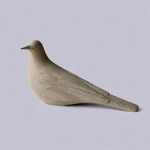 May include: A beige ceramic dove figurine in a resting position. The bird has detailed wing and body patterns. The dove's beak is pointed, and the tail is slightly elevated. The figurine is on a plain background.