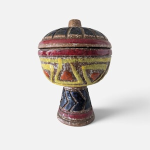Peut inclure: Un récipient en céramique avec couvercle et base sur pied, présentant une surface brune texturée avec des motifs géométriques peints à la main en rouge, jaune et bleu foncé. Le couvercle est bombé avec des rainures radiales.